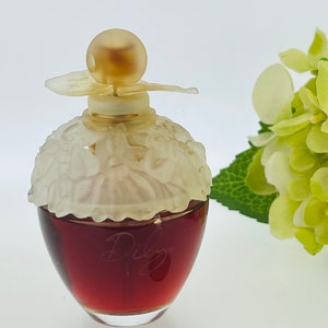 Może przedstawiać: Przezroczysta szklana butelka perfum z białą, kwiatową nakrętką i złotym wierzchołkiem. Butelka jest wypełniona ciemnoczerwonym płynem. Butelka stoi na białym tle.