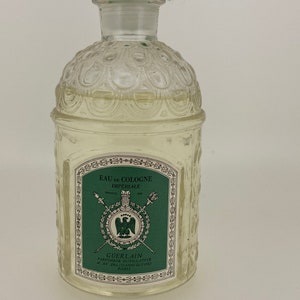 May include: A clear glass bottle of Guerlain Eau de Cologne Imperiale. The bottle has a textured design and a green label with gold lettering. The label reads "Eau de Cologne Imperiale" and "Guerlain Parfumeurs Distillateurs 67, Av. des Champs-Elysees Paris".