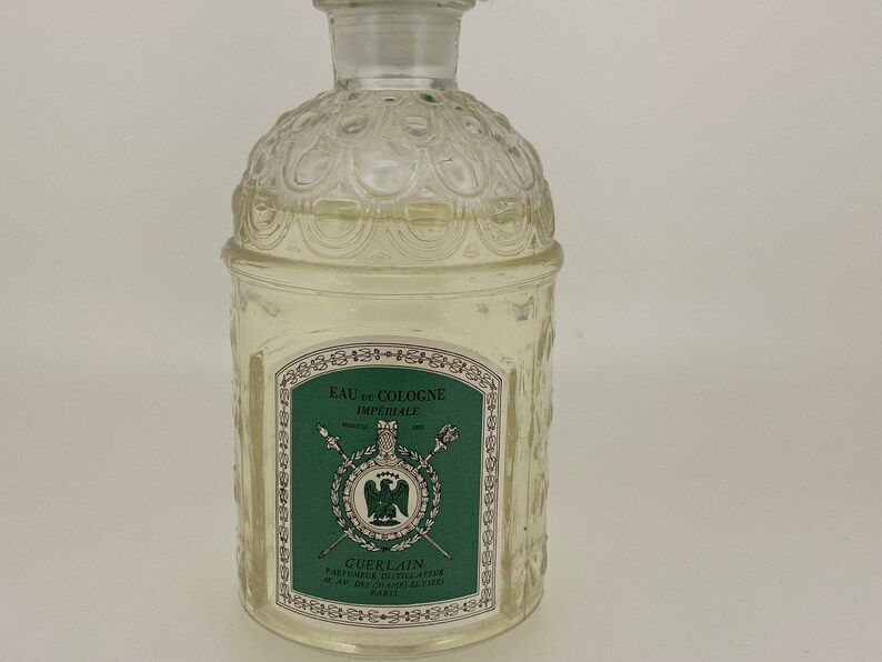 May include: A clear glass bottle of Guerlain Eau de Cologne Imperiale. The bottle has a textured design and a green label with gold lettering. The label reads "Eau de Cologne Imperiale" and "Guerlain Parfumeurs Distillateurs 67, Av. des Champs-Elysees Paris".