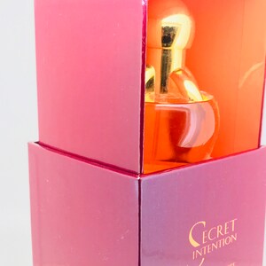 May include: A pink and orange box with a gold cap bottle of Secret Intention perfume inside. The bottle is clear and round with a gold cap.