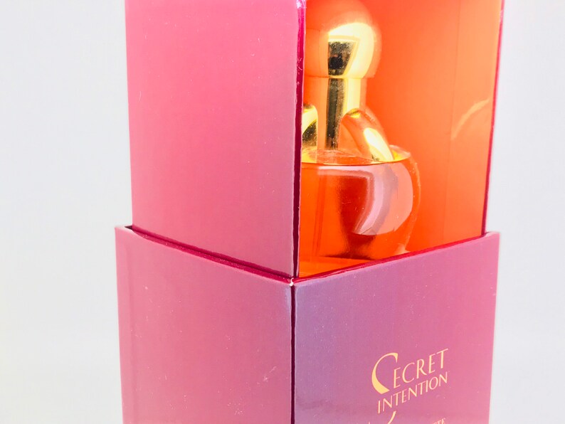May include: A pink and orange box with a gold cap bottle of Secret Intention perfume inside. The bottle is clear and round with a gold cap.