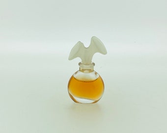 May include: A small, round perfume bottle with a clear glass body and a frosted white bird-shaped stopper. The bottle contains a light amber-colored liquid. The design is elegant and vintage, suitable for a perfume or fragrance.