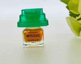 Può includere: Un piccolo flacone di profumo con un tappo verde a gradini. Il flacone contiene un liquido dorato e reca la scritta "Mitsouko Guerlain Paris". Su sfondo bianco, con fiori verdi e gialli.
