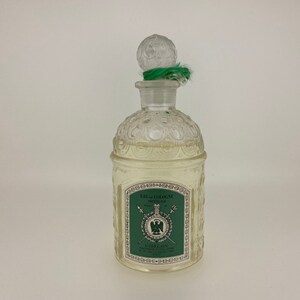 May include: A clear glass bottle of Guerlain Imperiale Eau de Cologne. The bottle has a textured design and a green stopper with a green ribbon. The label is green with gold lettering and features a crest with a green eagle.