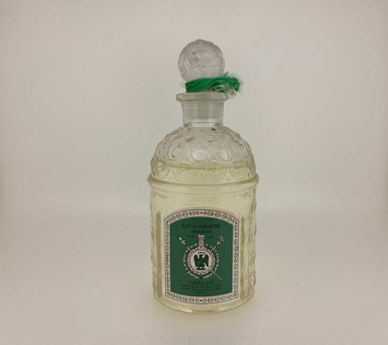 May include: A clear glass bottle of Guerlain Imperiale Eau de Cologne. The bottle has a textured design and a green stopper with a green ribbon. The label is green with gold lettering and features a crest with a green eagle.