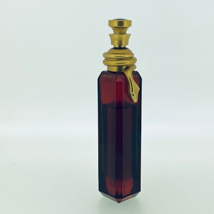 Op de afbeelding: Een donkerrode glazen fles met een gouden dop en een slangvormige stop. De fles is gevormd als een hoog, smal rechthoek met een puntige bodem.