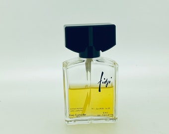 May include: A clear glass bottle of Fidji Eau de Toilette perfume by Guy Laroche. The bottle has a black cap and a black label with the word "Fidji" written in a stylized font.