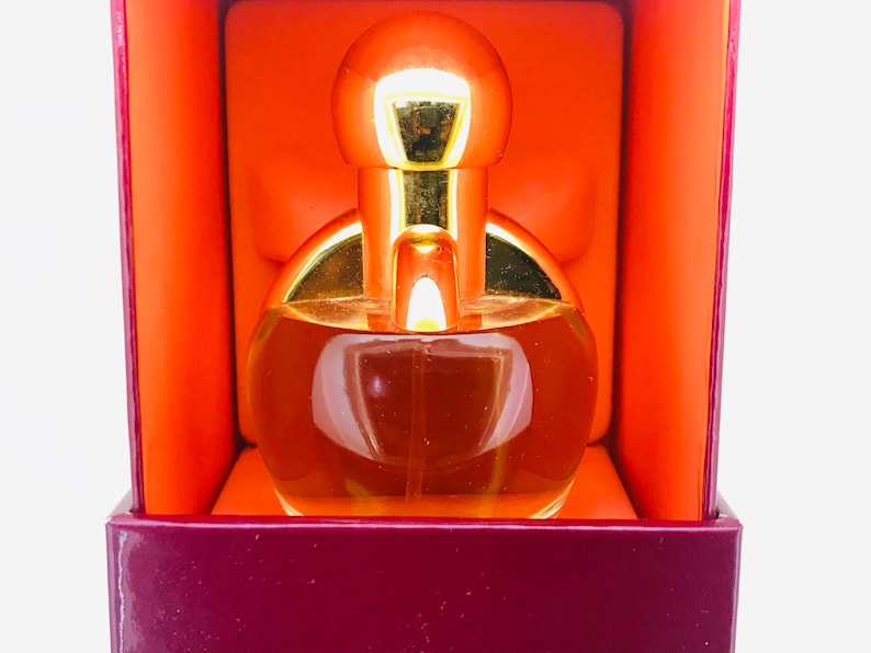 May include: A gold and clear glass perfume bottle with a round, bulbous top. The bottle is sitting inside a burgundy box with an orange interior.