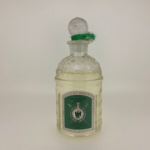 May include: A clear glass bottle of Guerlain Imperiale Eau de Cologne. The bottle has a textured design and a green stopper with a green ribbon. The label on the bottle is green and white and features the Guerlain logo and the text "Eau de Cologne Imperiale".