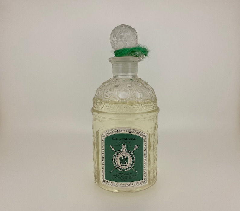 May include: A clear glass bottle of Guerlain Imperiale Eau de Cologne. The bottle has a textured design and a green stopper with a green ribbon. The label on the bottle is green and white and features the Guerlain logo and the text "Eau de Cologne Imperiale".