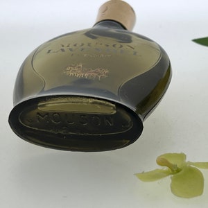 Op de afbeelding: Een vintage glazen flesje Mousson Lavandou parfum. De fles is donker amberkleurig en heeft een afgeronde vorm. Het etiket op de fles is goudkleurig en zegt "Mousson Lavandou" in een cursief schrift. De fles staat op een wit oppervlak.
