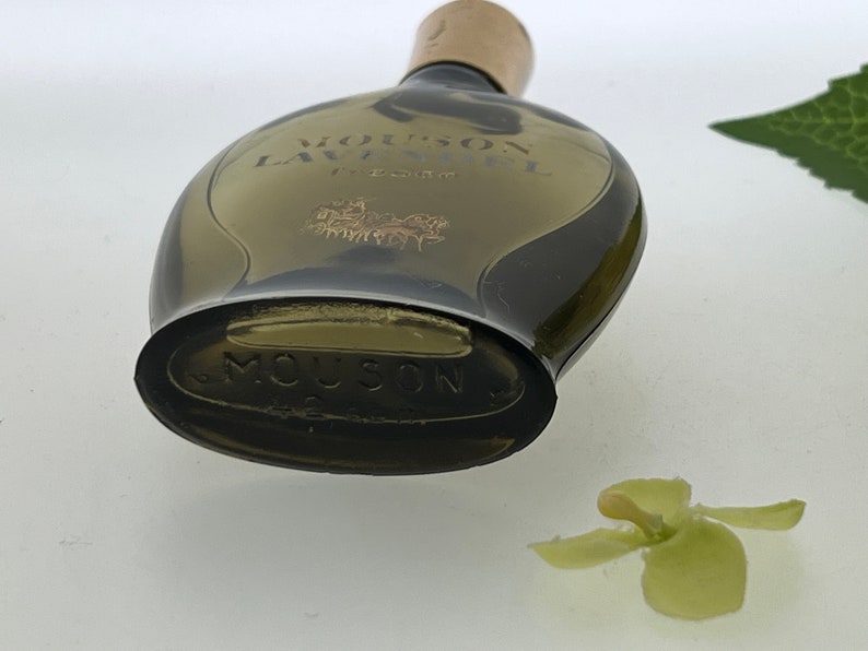 Op de afbeelding: Een vintage glazen flesje Mousson Lavandou parfum. De fles is donker amberkleurig en heeft een afgeronde vorm. Het etiket op de fles is goudkleurig en zegt "Mousson Lavandou" in een cursief schrift. De fles staat op een wit oppervlak.