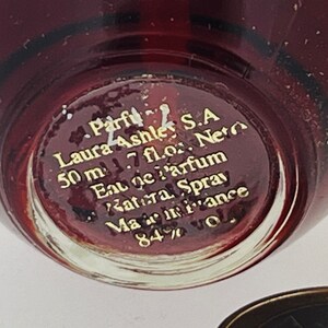 Może przedstawiać: Zbliżenie na sp&oacute;d szklanej butelki perfum Laura Ashley. Butelka jest czerwona i ma złote napisy: "Parfu Laura Ashley S.A 50 ml 1.7 fl.oz. Neto Eau De Parfum Katera Spray Mamiance 84%"