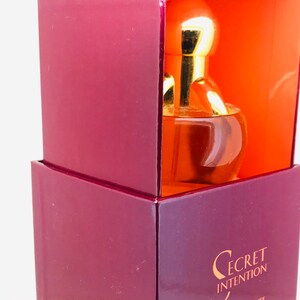 May include: A burgundy box with a gold lid and a clear glass bottle of perfume inside. The bottle has a gold cap and a gold ring around the neck. The box has the words "Secret Intention" printed on the front.