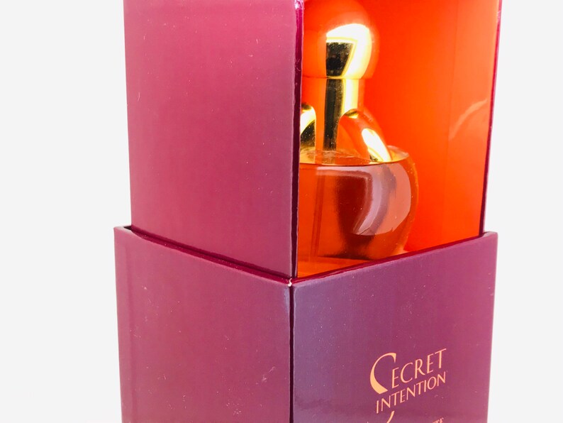 May include: A burgundy box with a gold lid and a clear glass bottle of perfume inside. The bottle has a gold cap and a gold ring around the neck. The box has the words "Secret Intention" printed on the front.
