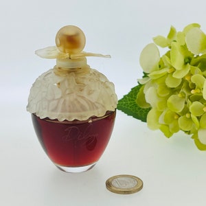 Może przedstawiać: Przezroczysta szklana butelka z białym, kwiatowym wzorem i złotą nakrętką. Butelka jest wypełniona ciemnoczerwonym płynem, a na przodzie wygrawerowane jest słowo "Dilis".