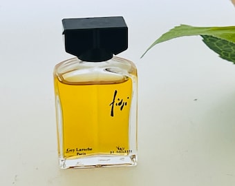 May include: A clear glass perfume bottle with a black square cap. The bottle contains a yellow liquid and has the word "fiji" in black script. The text "Guy Laroche Paris" is printed on the bottle.