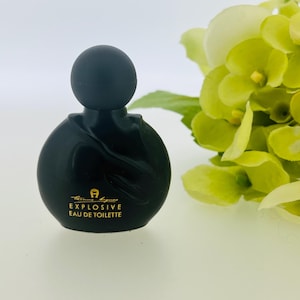 Puede incluir: Un frasco de perfume de vidrio negro con una etiqueta dorada que dice "Explosive Eau de Toilette".