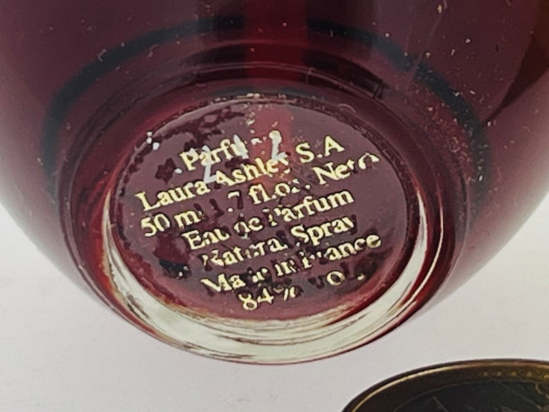 Może przedstawiać: Zbliżenie na sp&oacute;d szklanej butelki perfum Laura Ashley. Butelka jest czerwona i ma złote napisy: "Parfu Laura Ashley S.A 50 ml 1.7 fl.oz. Neto Eau De Parfum Katera Spray Mamiance 84%"