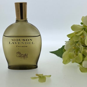 Op de afbeelding: Een bruine glazen fles Mousson LavendeL Fresco cologne met een gouden dop. De fles staat op een wit oppervlak met een groene bloem op de voorgrond.