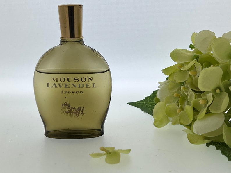 Op de afbeelding: Een bruine glazen fles Mousson LavendeL Fresco cologne met een gouden dop. De fles staat op een wit oppervlak met een groene bloem op de voorgrond.
