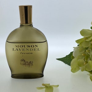 Op de afbeelding: Een bruine glazen fles Mousson LavendeL fresco cologne met een gouden dop. De fles staat op een wit oppervlak met een groene bloem op de achtergrond.