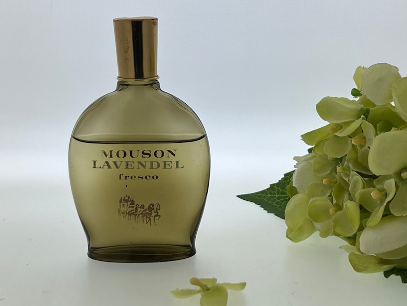 Op de afbeelding: Een bruine glazen fles Mousson LavendeL fresco cologne met een gouden dop. De fles staat op een wit oppervlak met een groene bloem op de achtergrond.
