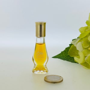 May include: A small, clear glass bottle of perfume with a gold cap. The bottle is shaped like a teardrop and has a label that says "Cacharel" in white lettering. The bottle is sitting on a white surface next to a gold coin.