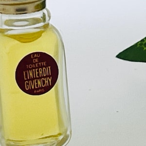 Puede incluir: Un pequeño frasco de perfume de vidrio transparente con una tapa dorada. La etiqueta dice "Eau de Toilette L'Interdit Givenchy Paris".