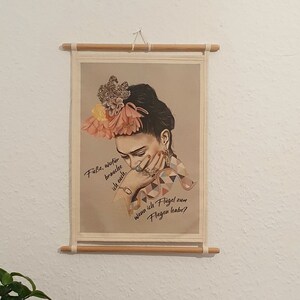 Può includere: Una stampa incorniciata di una donna con una corona di fiori colorati sulla testa. La donna sta piangendo e il testo sulla stampa dice "Falsche Träume brauchen dich nicht, ich weiß, ich flieg' zum Mond, Frag' den Mond!"