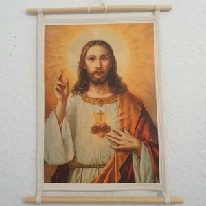 May include: A vintage-style religious print of Jesus Christ with a golden halo and a red heart with thorns on his chest. The print is framed with a wooden frame and hung on a white wall.