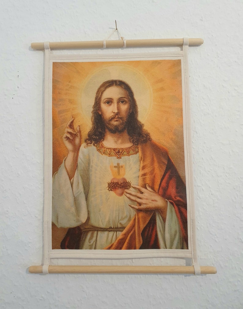 May include: A vintage-style religious print of Jesus Christ with a golden halo and a red heart with thorns on his chest. The print is framed with a wooden frame and hung on a white wall.