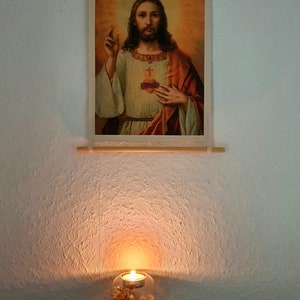 May include: A framed image of Jesus Christ with a heart on his chest, hanging on a white wall. A glass candle holder with dried flowers and a lit candle sits on a black surface below the image.