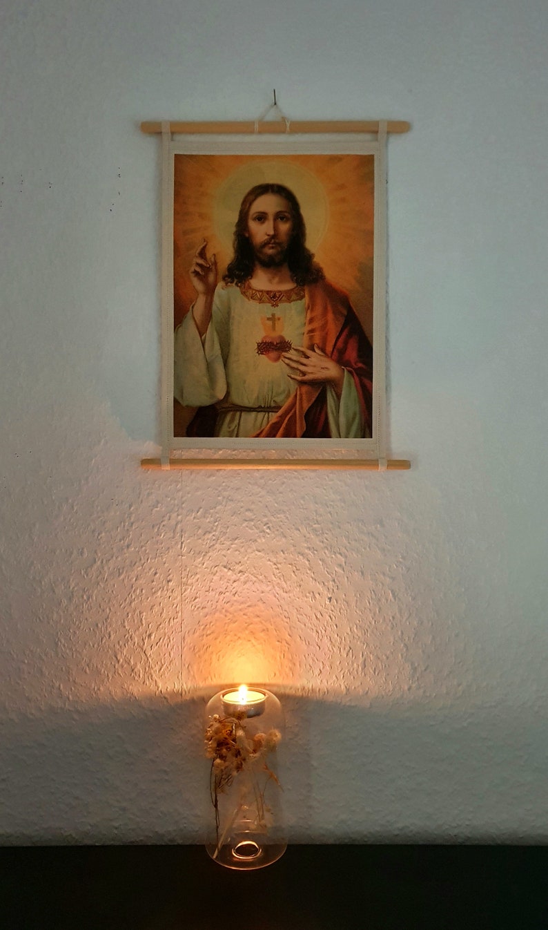 May include: A framed image of Jesus Christ with a heart on his chest, hanging on a white wall. A glass candle holder with dried flowers and a lit candle sits on a black surface below the image.
