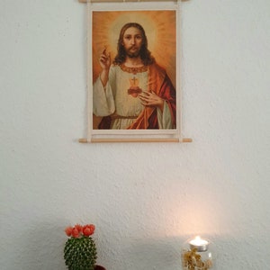 May include: A framed religious print of Jesus Christ with a golden halo and a red and gold robe. The print is hanging on a white wall with a cactus plant and a candle holder in the foreground.