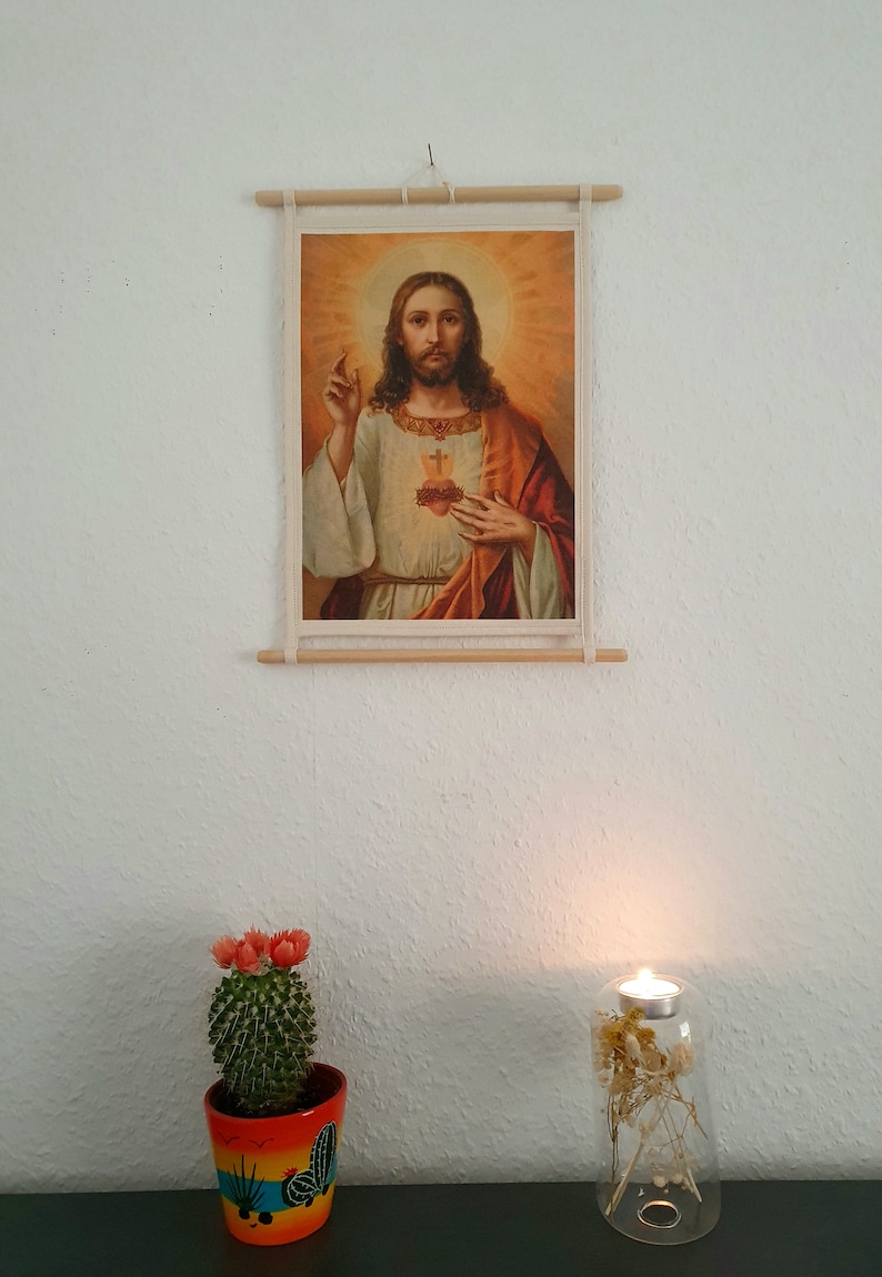 May include: A framed religious print of Jesus Christ with a golden halo and a red and gold robe. The print is hanging on a white wall with a cactus plant and a candle holder in the foreground.