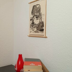 May include: A black and white illustration of a creature with a human-like face and a tree-like body. The illustration is printed on a canvas and framed with a wooden dowel. The canvas is hanging on a white wall.
