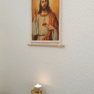 May include: A framed print of a religious image depicting Jesus Christ with a heart on his chest. The print is hanging on a white wall with a lit candle in a glass jar with dried flowers below it.