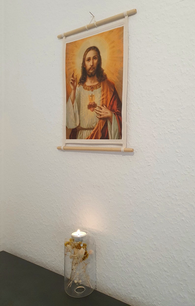 May include: A framed print of a religious image depicting Jesus Christ with a heart on his chest. The print is hanging on a white wall with a lit candle in a glass jar with dried flowers below it.