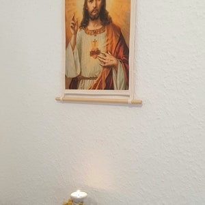 May include: A framed canvas print of a painting depicting Jesus Christ with a heart on his chest. The print is hanging on a white wall. A glass candle holder with dried flowers is on a dark surface below the print.