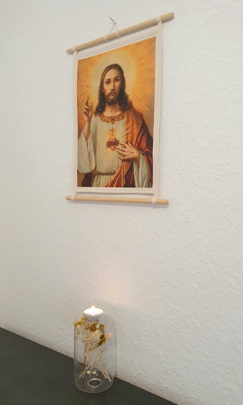 May include: A framed canvas print of a painting depicting Jesus Christ with a heart on his chest. The print is hanging on a white wall. A glass candle holder with dried flowers is on a dark surface below the print.