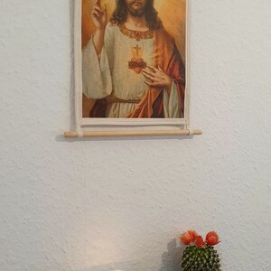 May include: A framed religious print of Jesus Christ with a heart on his chest, a cactus in a colourful pot, and a glass candle holder with dried flowers.