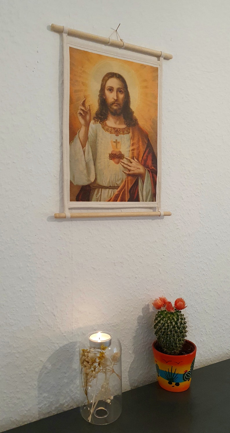 May include: A framed religious print of Jesus Christ with a heart on his chest, a cactus in a colourful pot, and a glass candle holder with dried flowers.
