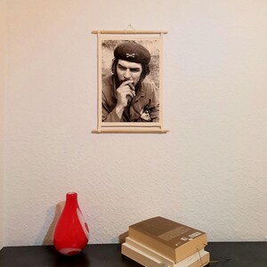 May include: A wall-mounted scroll featuring a black and white photographic portrait of a man wearing a beret and smoking a cigar. The print is held by light-colored wooden rods at the top and bottom, suspended by a thin string from the top rod.