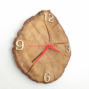 Könnte beinhalten: Eine rustikale Wanduhr aus einem Baumstammabschnitt. Das Zifferblatt zeigt natürliche Holzmaserung mit goldfarbenen Ziffern bei 3, 6, 9 und 12. Rote Zeiger zeigen die Uhrzeit an. Die Uhr ist an einer weißen Wand befestigt.