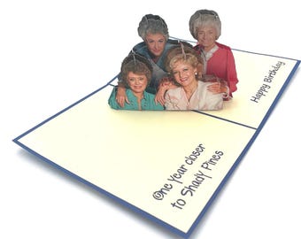 May include: A pop-up birthday card featuring four women with text that reads "Happy Birthday" and "One year closer to Shady Pines". The card has a cream-coloured background with a blue border.