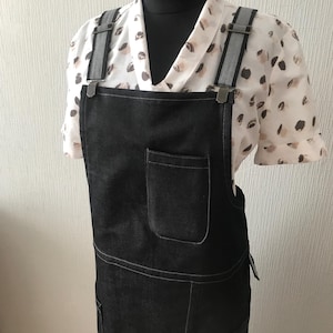 May include: Black denim apron with adjustable straps and two pockets. The apron is worn over a white blouse with a brown and white pattern.