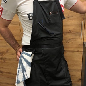 Könnte beinhalten: Eine schwarze Schürze mit einer großen Tasche auf der Vorderseite, einer kleineren Tasche an der Seite und einem Bindegürtel. Die Schürze wird über einem weißen Hemd mit roten und weißen Streifen und dem Wort "Bayern" auf der Vorderseite getragen.