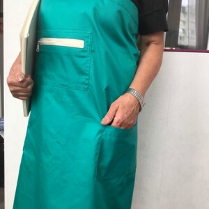 Puede incluir: Un delantal verde azulado con tirantes blancos y un bolsillo blanco con cremallera. El delantal tiene un bolsillo grande en el lado derecho.