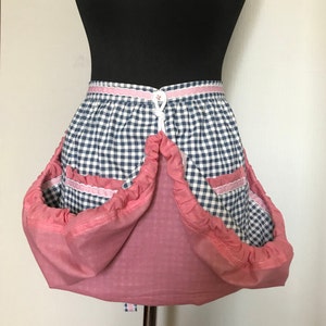 May include: A pink and blue gingham apron with large pockets. The apron has a white ribbon tie and a pink trim.